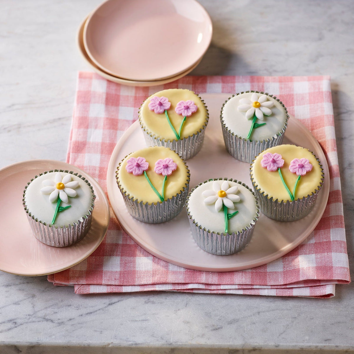 Fiona Cairns Floral Golden Fairy Cakes with smooth fondant icing and handcrafted sugar paste daisy and flower decorations, arranged on a pink plate with a gingham cloth on a marble surface.