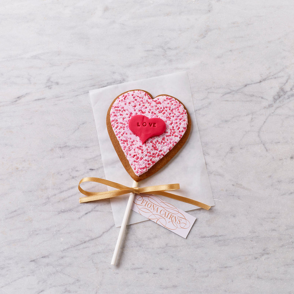 Heart-shaped iced gingerbread on a stick with pink sprinkles and a red “love” heart, tied with a ribbon and styled on a marble surface.