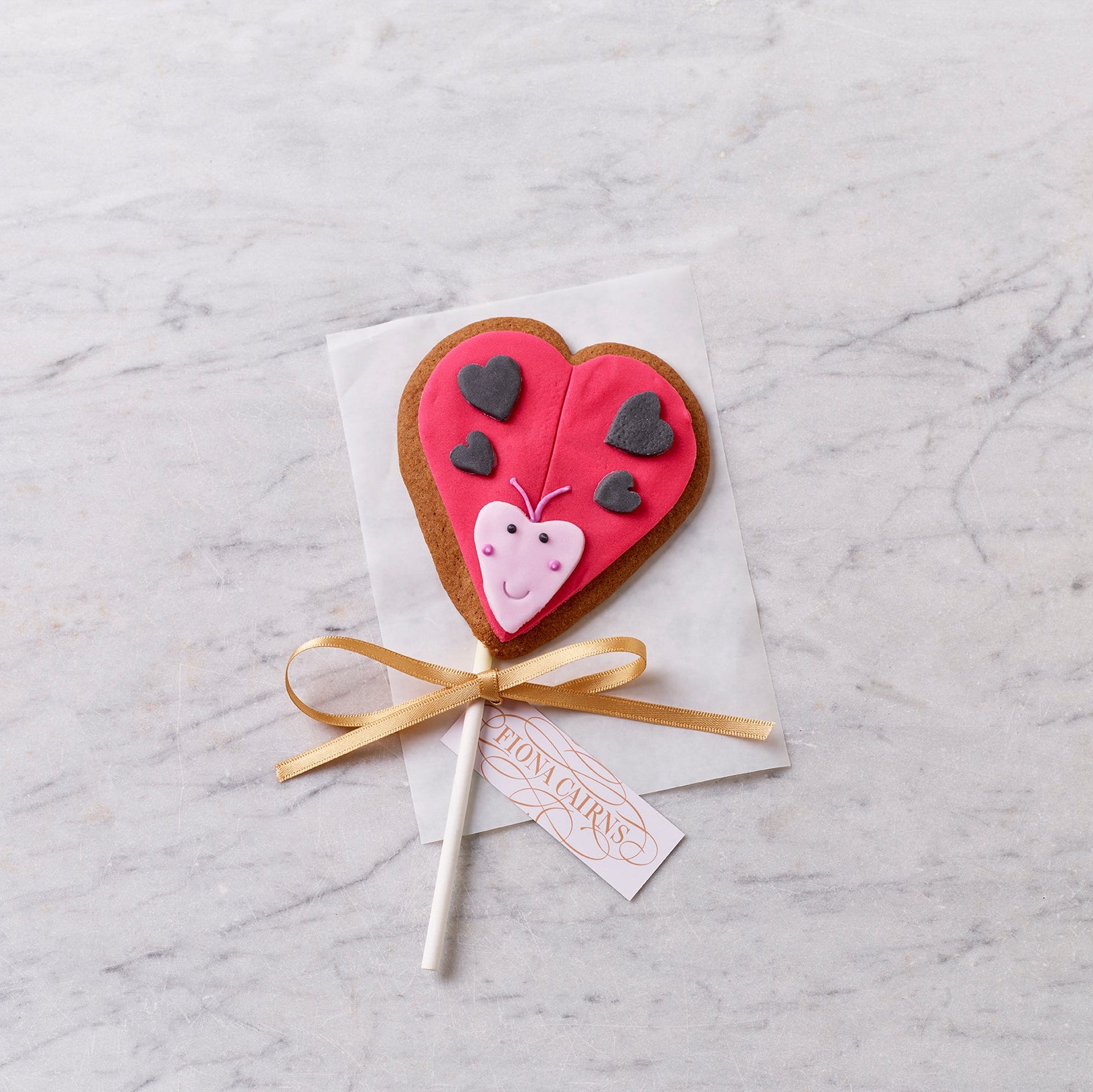 Heart-shaped decorated cookie on a stick with pink icing, butterfly detail and ribbon, styled on a marble background.