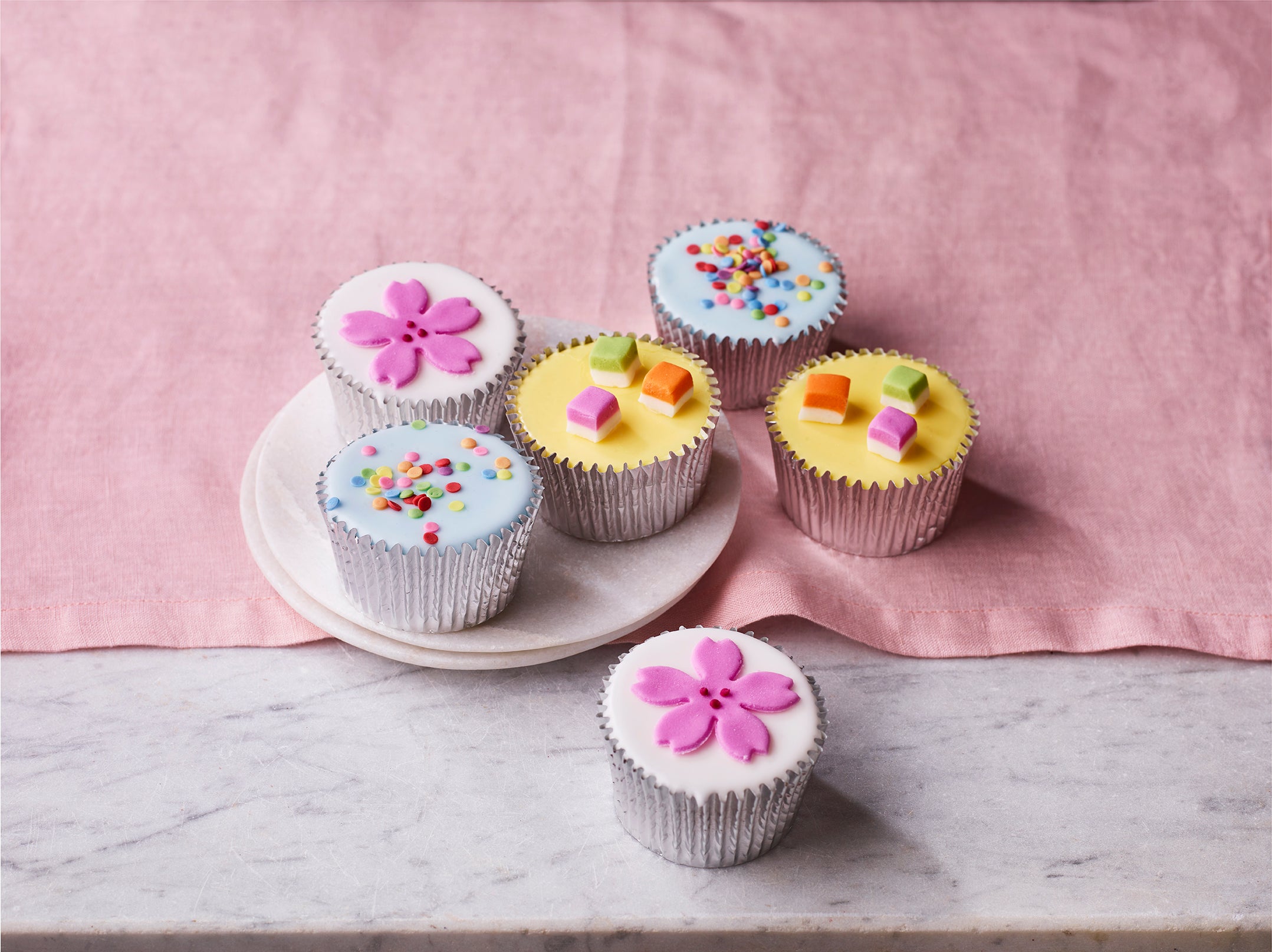 Confetti fairy cakes with colourful icing decorations and sprinkles in silver cases on a plate with a pink cloth background