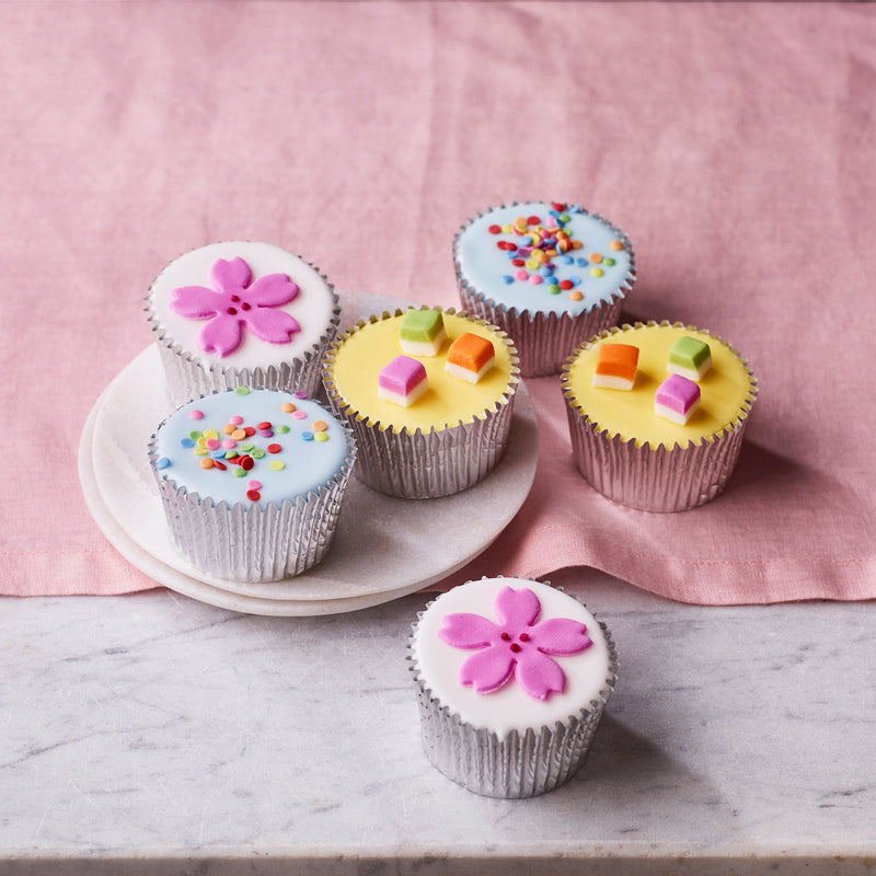 Confetti fairy cakes with colourful icing decorations and sprinkles in silver cases on a plate with a pink cloth background