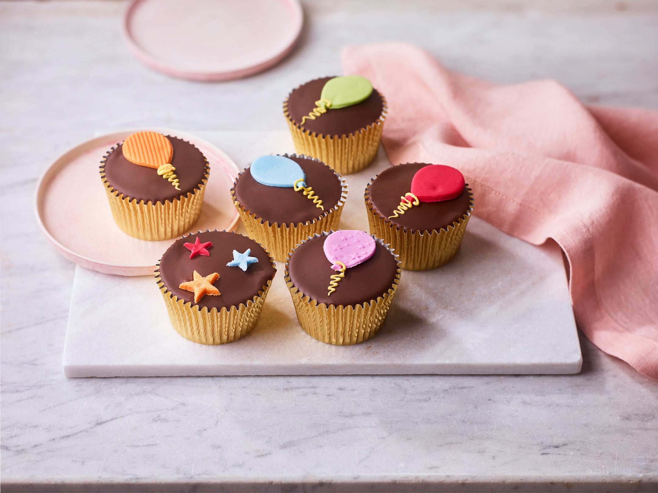 Six chocolate fairy cakes topped with smooth chocolate fondant and colourful sugar paste balloon and star decorations, arranged on a marble board with a pink napkin.