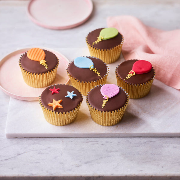Six chocolate fairy cakes topped with smooth chocolate fondant and colourful sugar paste balloon and star decorations, arranged on a marble board with a pink napkin.