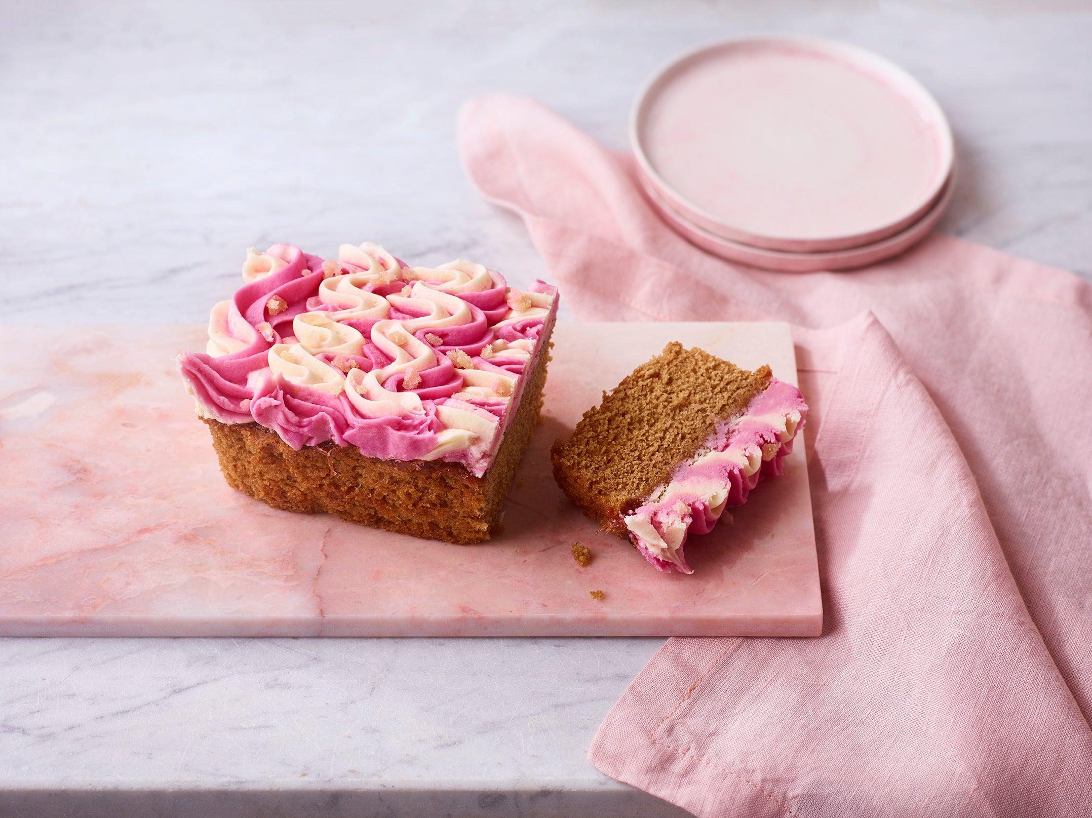 Rhubarb and ginger loaf cake with pink and white swirled frosting, sliced on a marble board with a pink napkin and plates in the background.