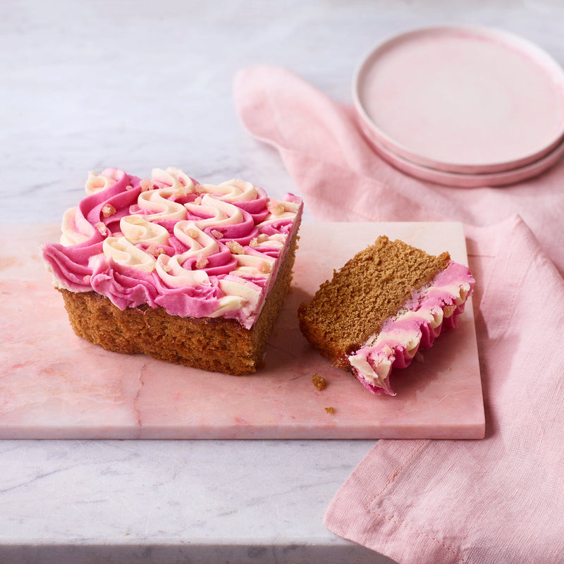 Rhubarb and ginger loaf cake with pink and white swirled frosting, sliced on a marble board with a pink napkin and plates in the background.