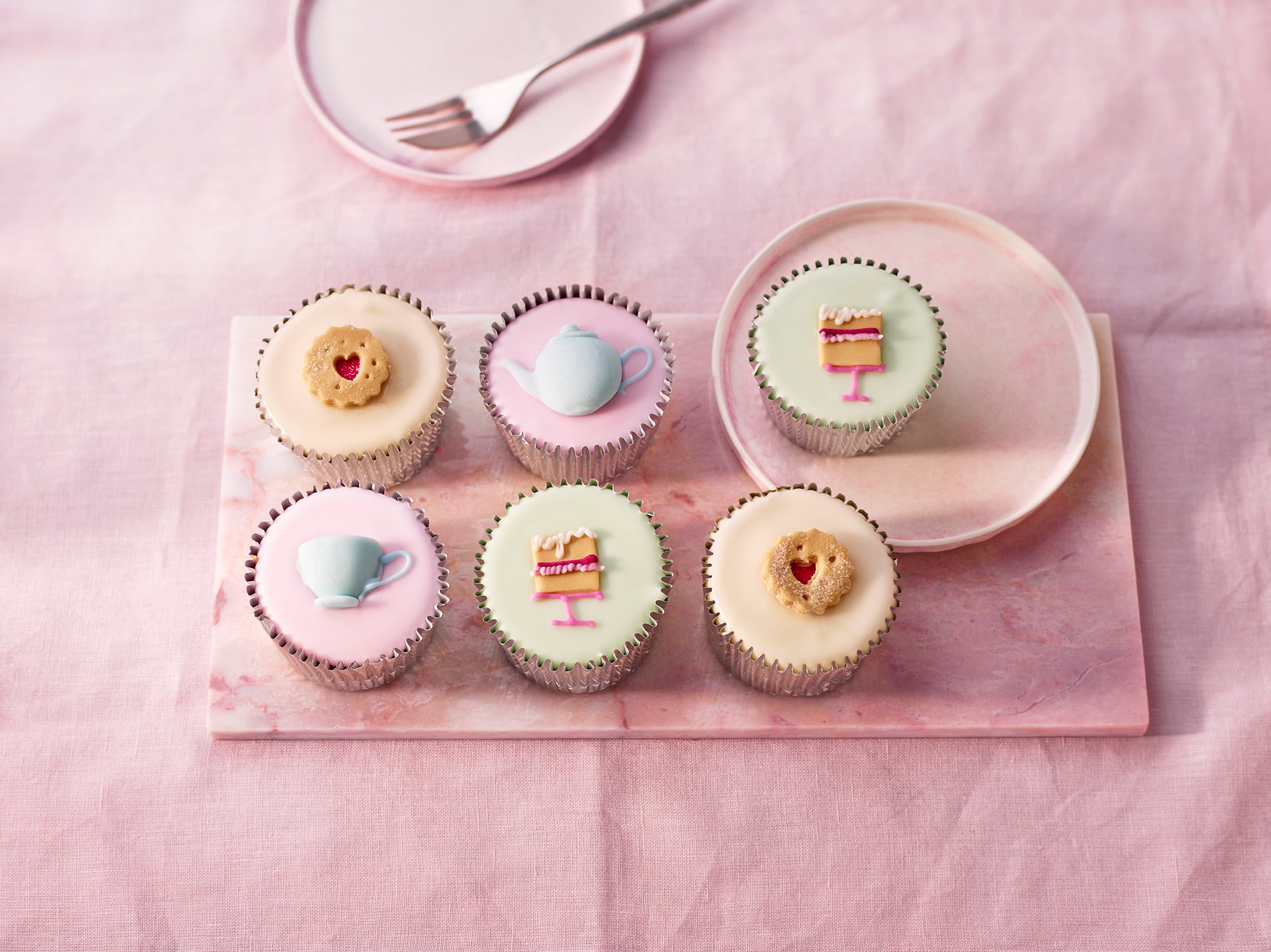 Afternoon Tea Fairy Cakes, six pastel fondant cakes with handcrafted teacup, teapot, cake slice and heart biscuit sugar decorations on a pink board.