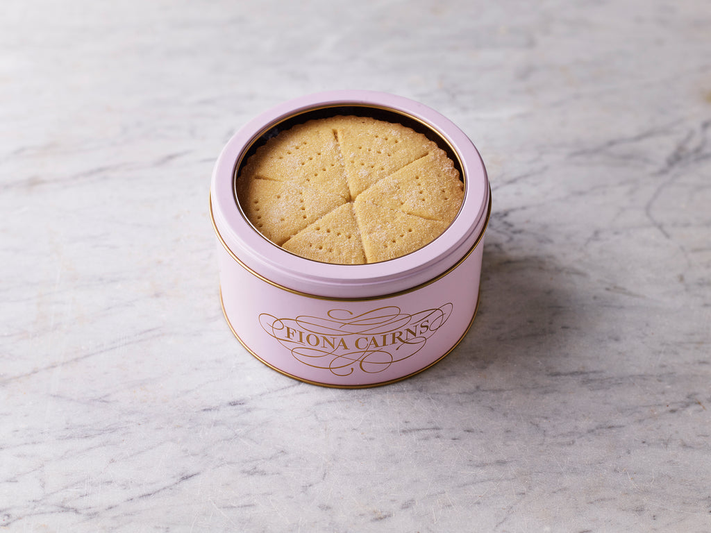 Round pink tin labelled “Fiona Cairns” filled with shortbread wedges, sitting on a light marble surface.