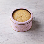 Round pink tin labelled “Fiona Cairns” filled with shortbread wedges, sitting on a light marble surface.
