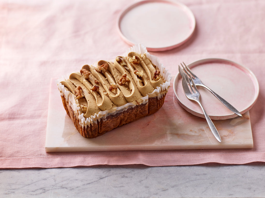 Coffee, walnut and cardamom loaf cake with piped coffee frosting and walnut pieces on a marble board with pink plates and cutlery in the background.