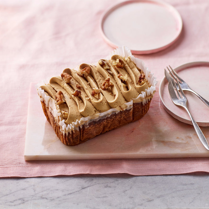 Coffee, walnut and cardamom loaf cake with piped coffee frosting and walnut pieces on a marble board with pink plates and cutlery in the background.