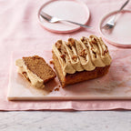 Sliced coffee, walnut and cardamom loaf cake with piped coffee frosting and walnut pieces on a marble board with pink plates and cutlery in the background.