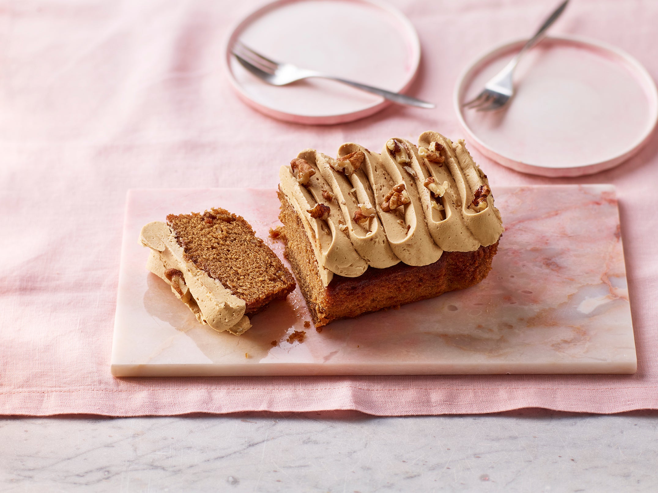 Sliced coffee, walnut and cardamom loaf cake with piped coffee frosting and walnut pieces on a marble board with pink plates and cutlery in the background.