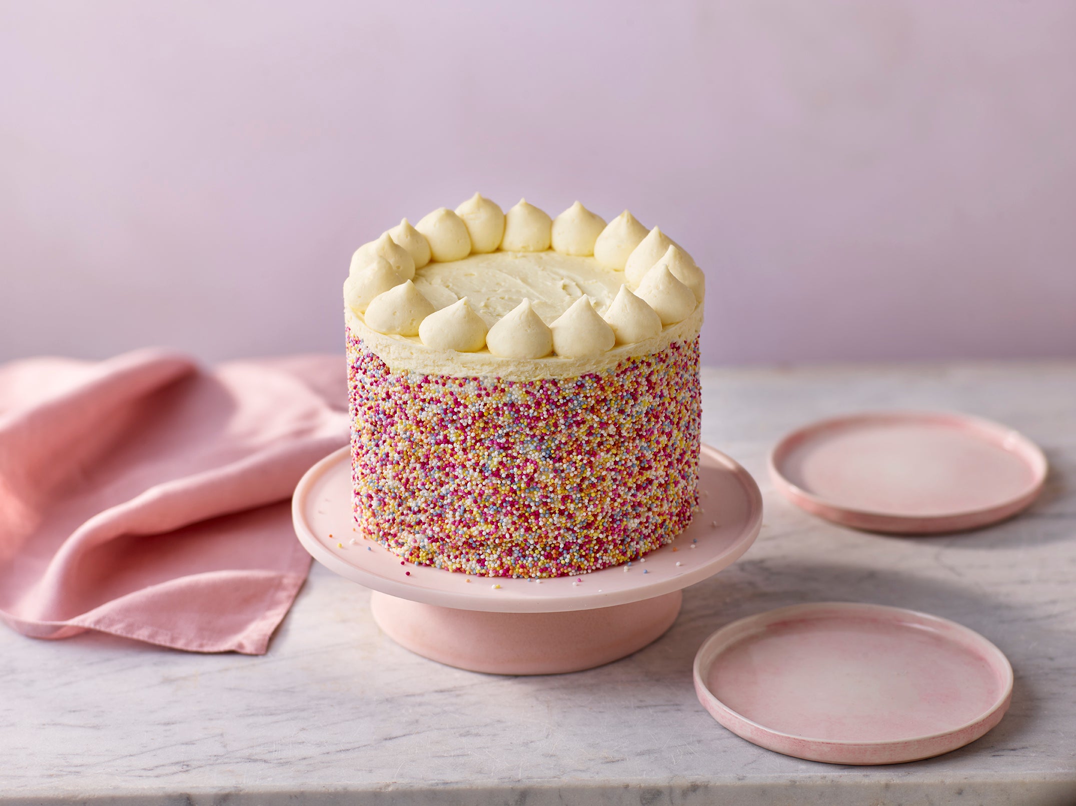 Colorful cake with white frosting on a pink stand against a light purple background