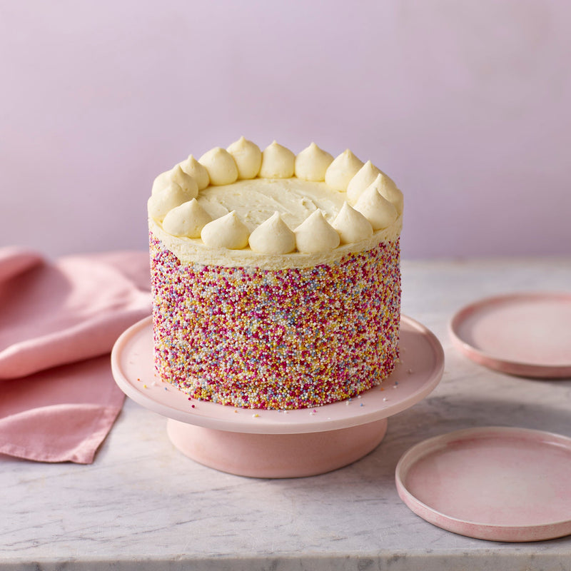 Colorful cake with white frosting on a pink stand against a light purple background