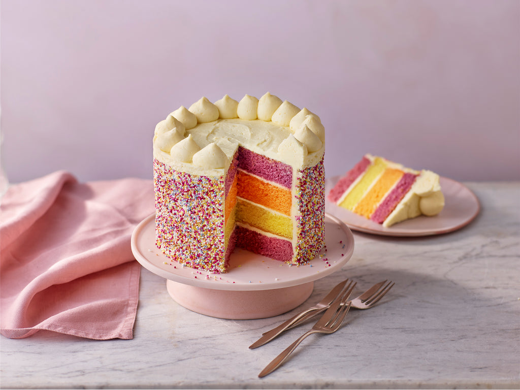 Colorful layered cake with a slice cut out on a pink cake stand, placed on a light wooden surface.