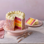 Colorful layered cake with a slice cut out on a pink cake stand, placed on a light wooden surface.