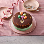 6-inch chocolate Happy Birthday cake with balloon decoration on a pink plate and ribbon background.