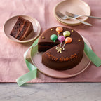 6-inch chocolate Happy Birthday cake sliced with balloon decoration on a pink plate and ribbon background.