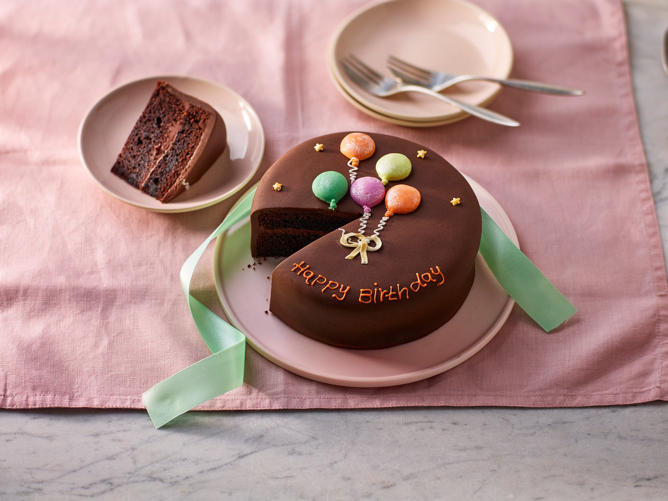 6-inch chocolate Happy Birthday cake sliced with balloon decoration on a pink plate and ribbon background.