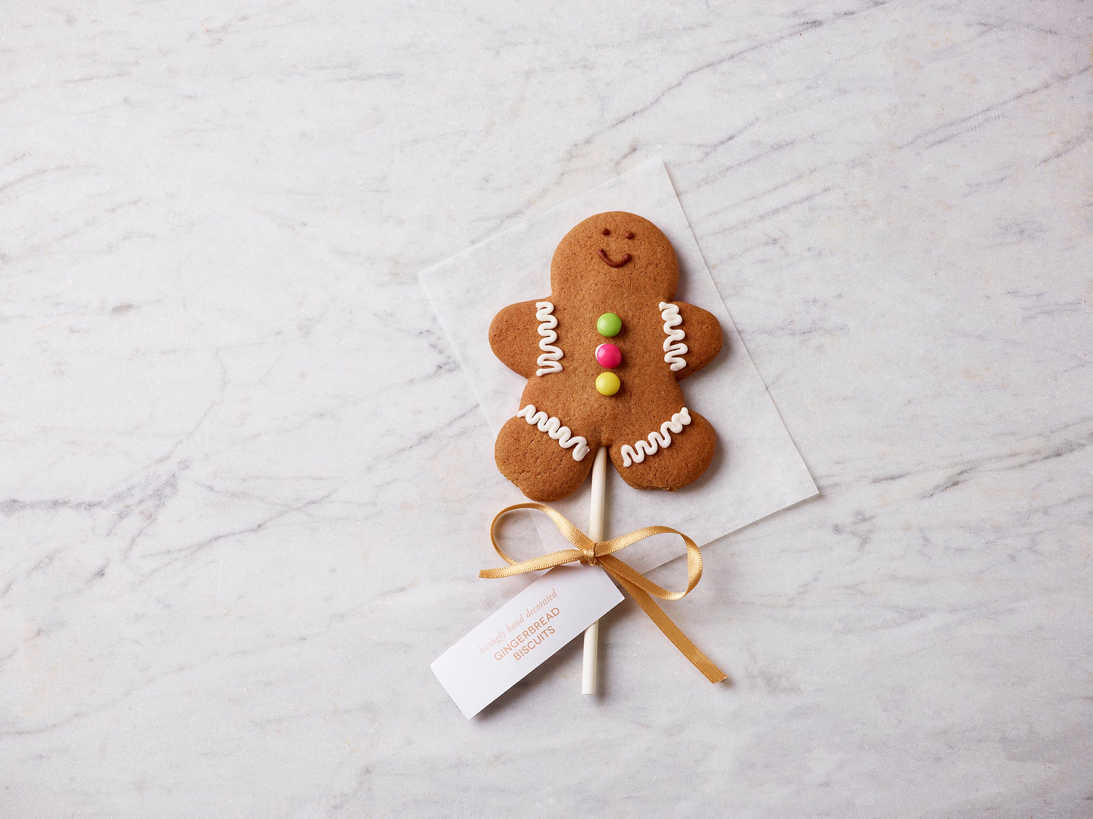 Gingerbread man lollipop decorated with icing and colourful buttons, tied with a gold ribbon and displayed on a marble background.