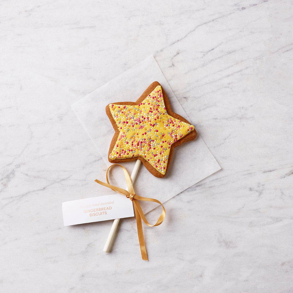 Hand-decorated gingerbread star biscuit lollipop with yellow icing, sprinkles and gold ribbon, photographed on a light marble surface