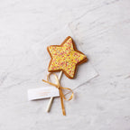 Hand-decorated gingerbread star biscuit lollipop with yellow icing, sprinkles and gold ribbon, photographed on a light marble surface