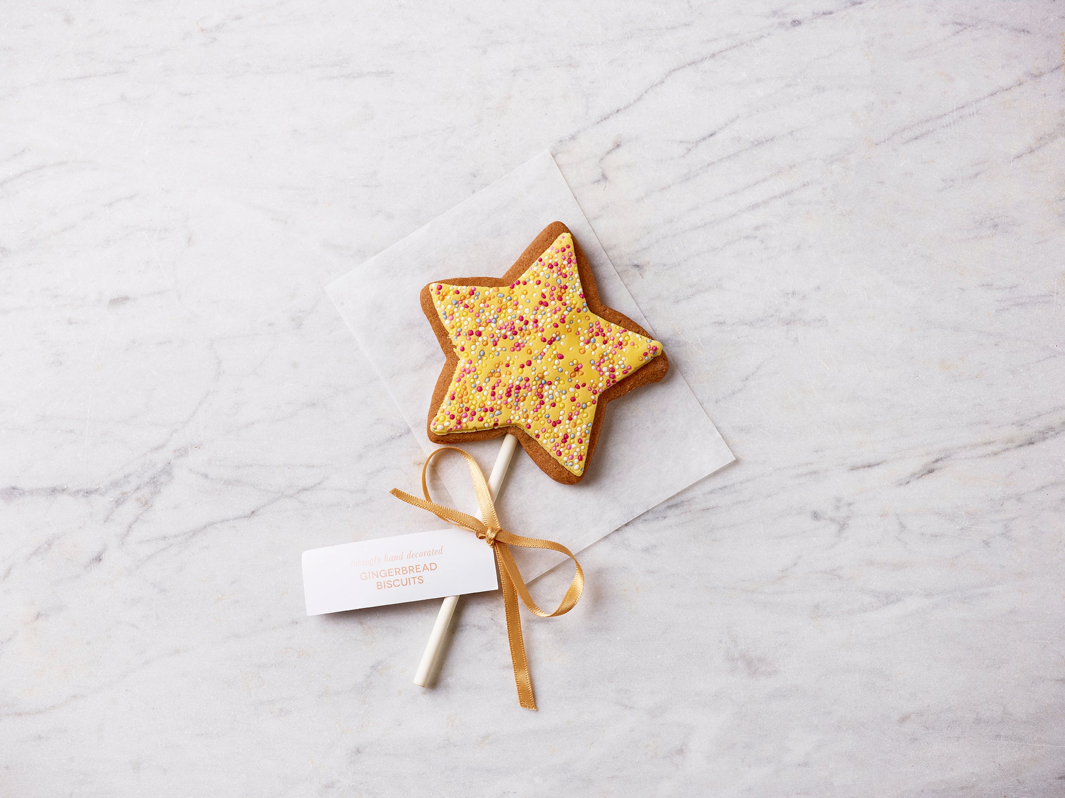 Hand-decorated gingerbread star biscuit lollipop with yellow icing, sprinkles and gold ribbon, photographed on a light marble surface