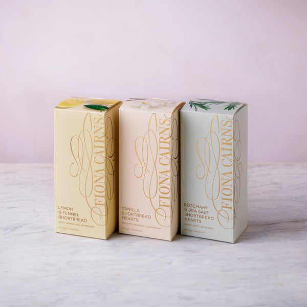 Biscuit and teatime treats bundle featuring three boxes of Fiona Cairns shortbread in pastel packaging, displayed on a marble surface with a soft pink background.