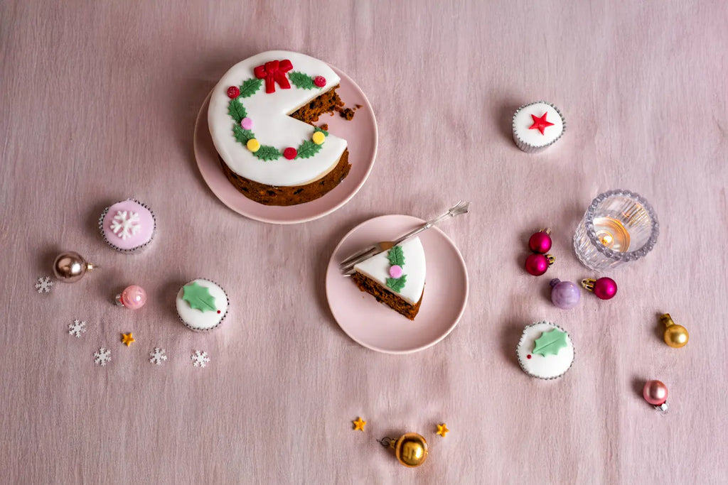 Top iced Christmas holly wreath fruitcake with a slice served on a pink plate, surrounded by mini decorated cakes and Christmas ornaments on a pink tablecloth.