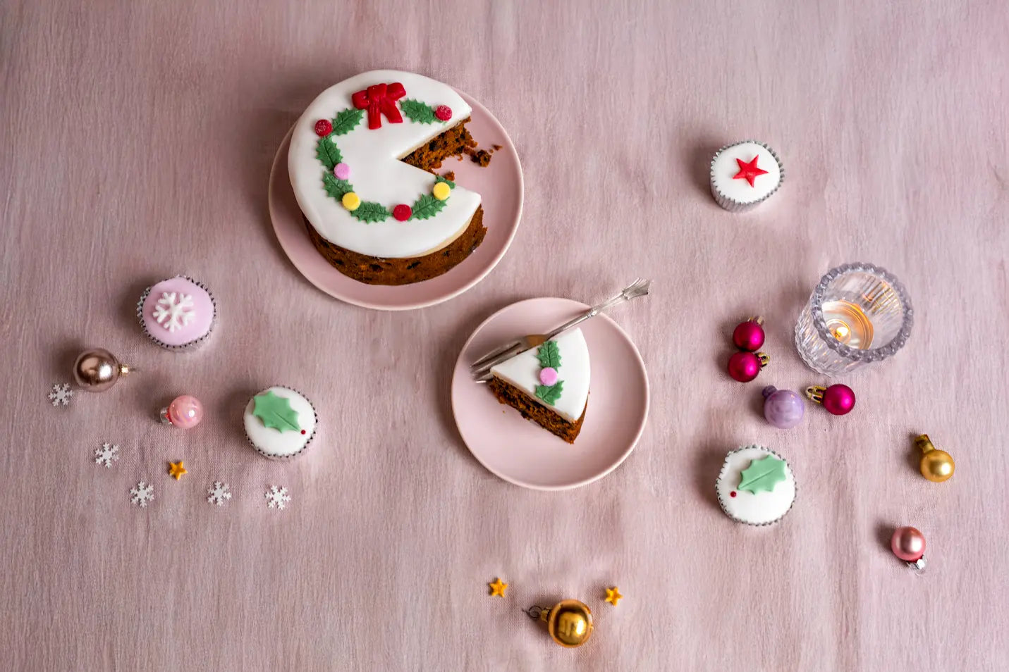 Top iced Christmas holly wreath fruitcake with a slice served on a pink plate, surrounded by mini decorated cakes and Christmas ornaments on a pink tablecloth.