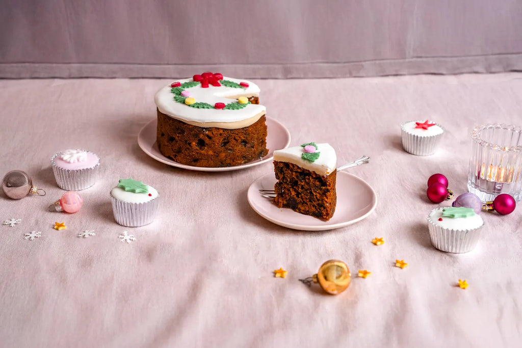 5-inch top iced Christmas holly wreath fruitcake with a slice served on a pink plate, surrounded by mini Christmas cakes and festive ornaments on a soft pink background.