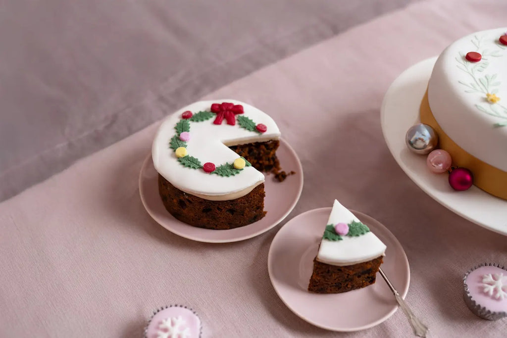 5-inch top iced Christmas holly wreath fruitcake with a slice on a pink plate, styled with festive ornaments and mini Christmas cakes on a soft pink background.