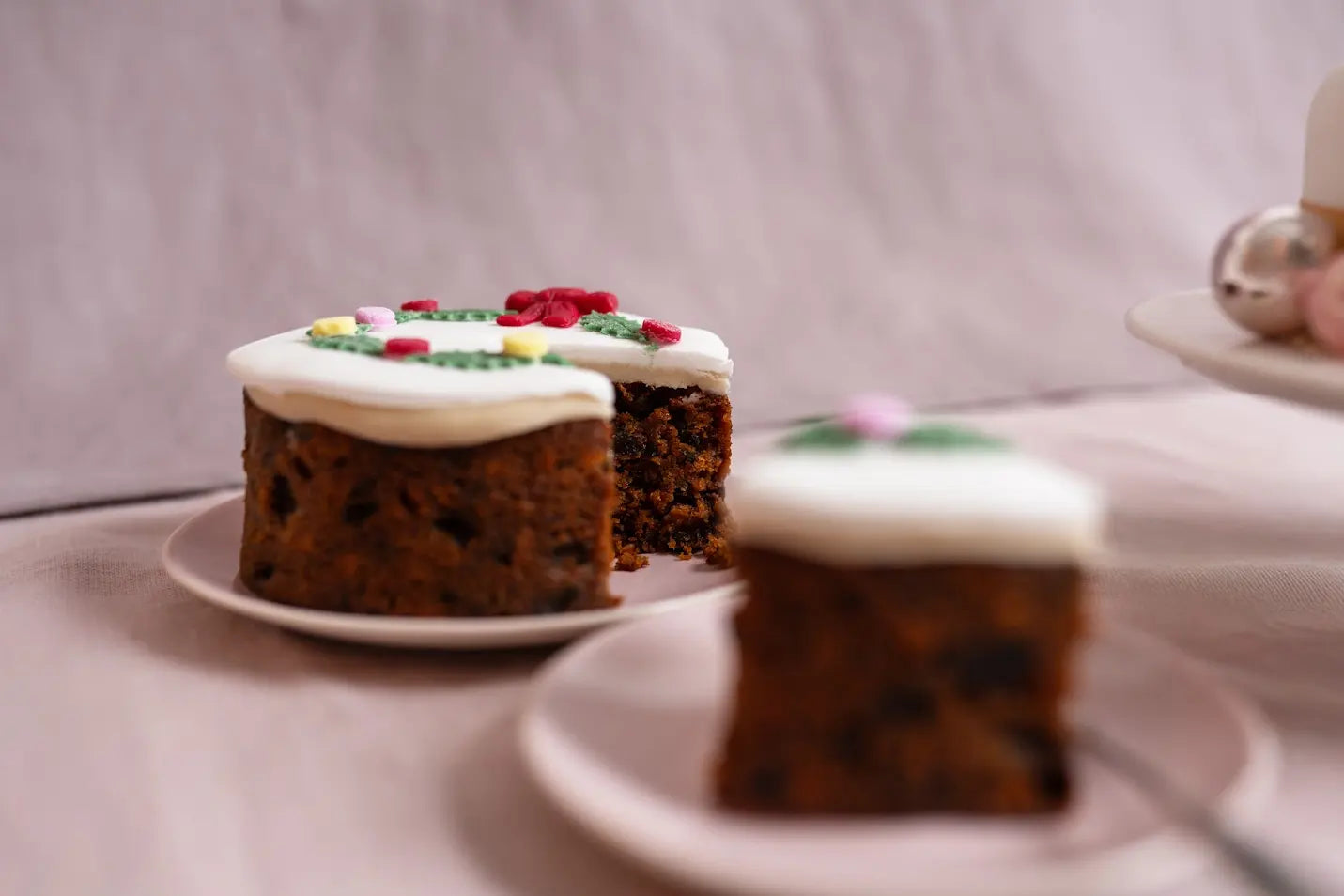 Close-up of a 5-inch top iced Christmas holly wreath fruitcake with a slice served on a pink plate against a soft pink background