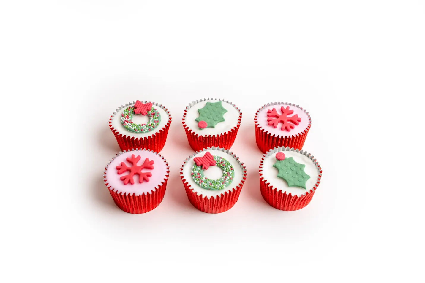 Christmas wreath fairy cakes decorated with holly leaves, snowflakes, wreaths and sprinkles in red cupcake cases, arranged on a white background.