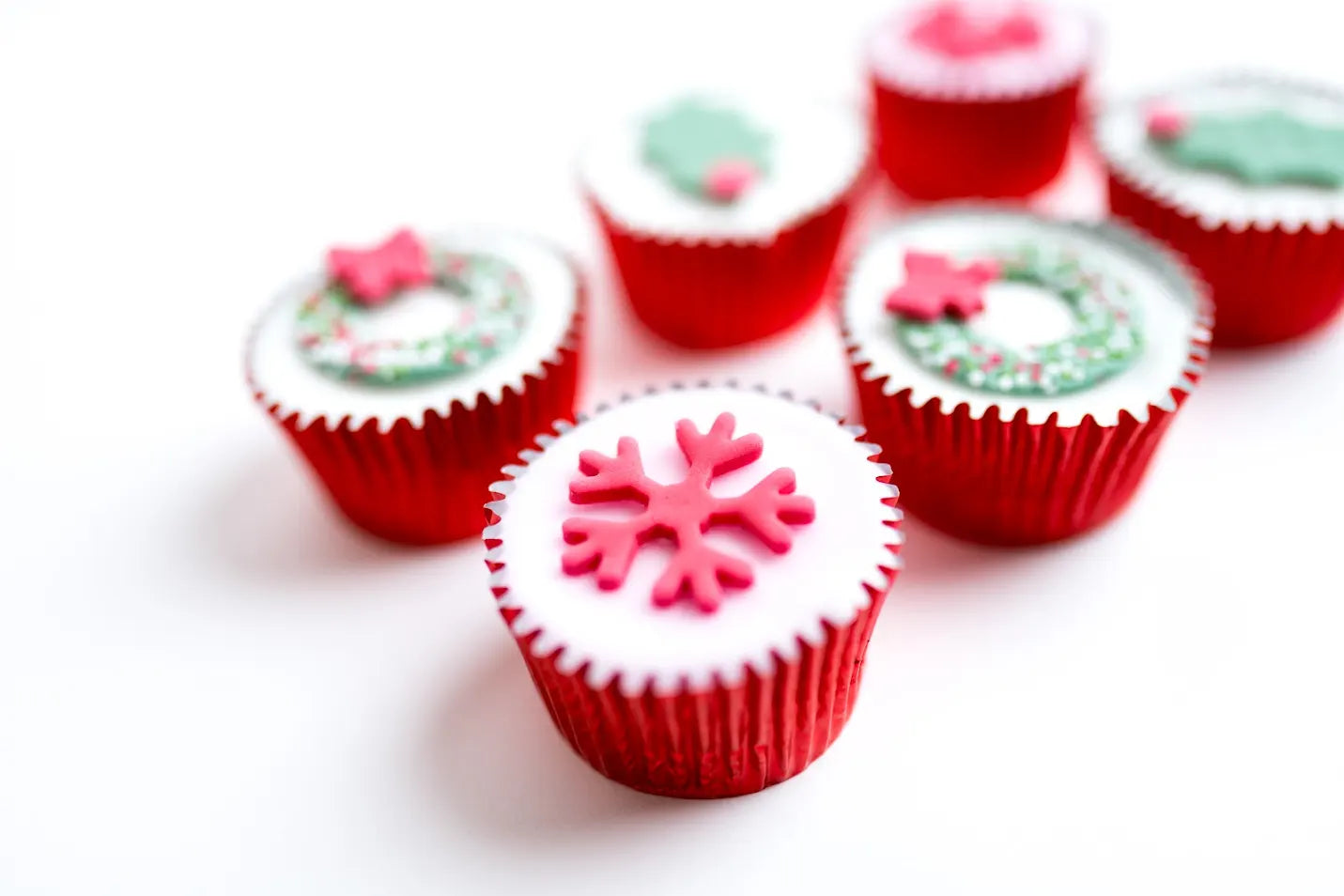 Christmas wreath fairy cakes decorated with holly leaves, snowflakes, wreaths and sprinkles in red cupcake cases, arranged on a white background.