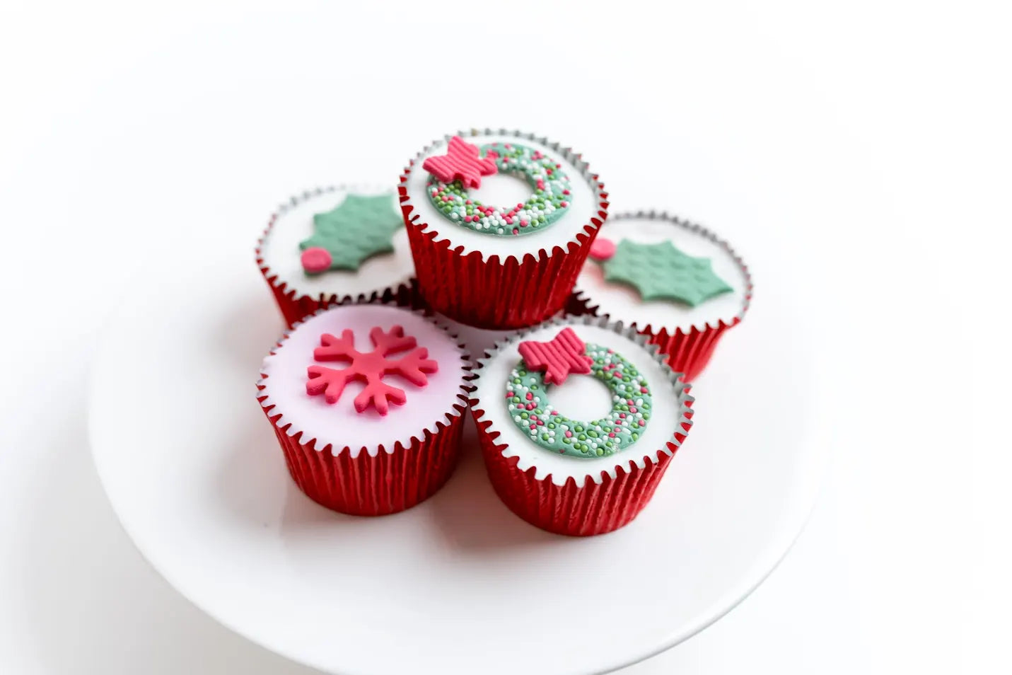 Christmas wreath fairy cakes decorated with holly leaves, snowflake, wreaths and sprinkles in red cupcake cases, arranged on a white plate.