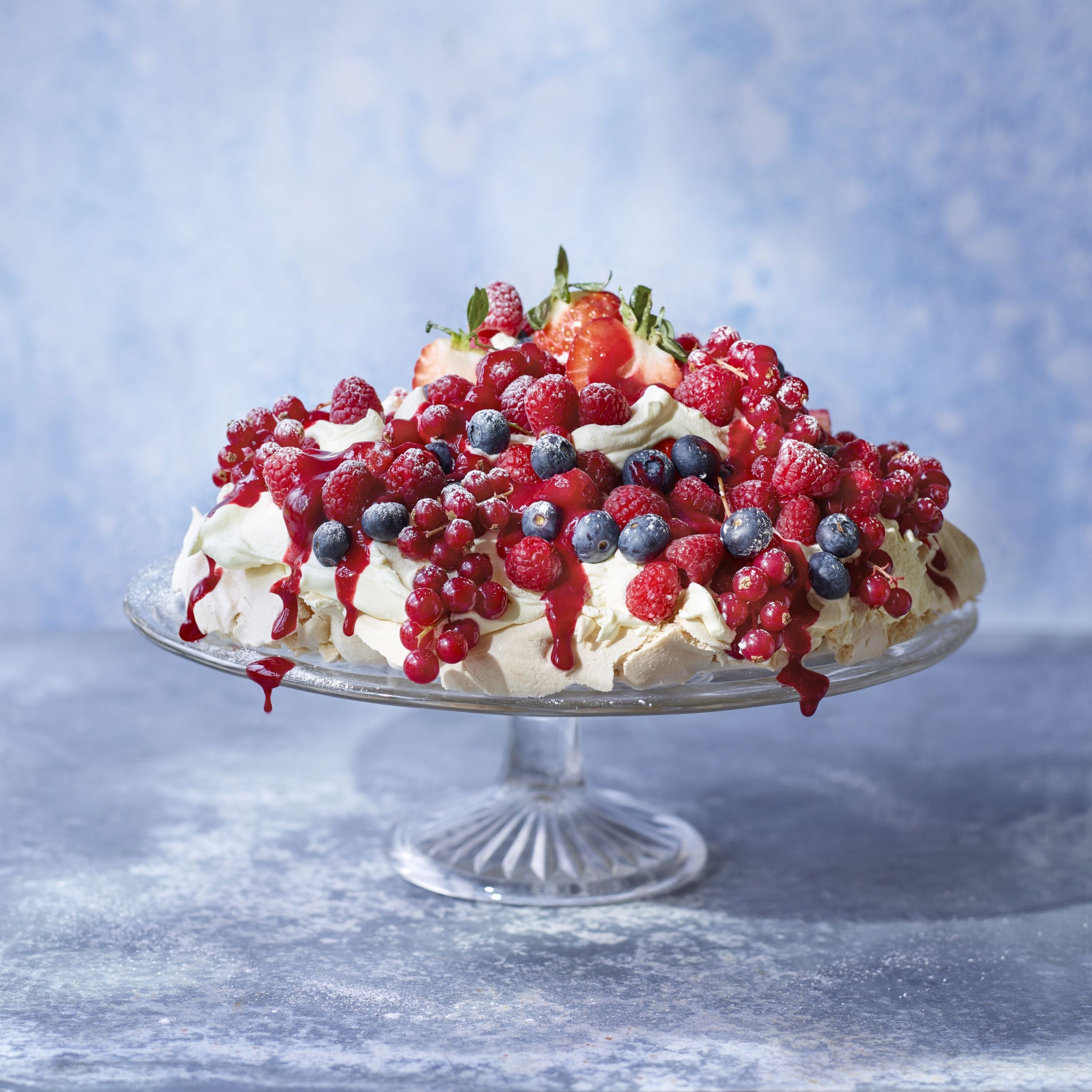 Berry pavlova dessert topped with fresh strawberries, raspberries, redcurrants and blueberries on a glass cake stand against a textured blue background.