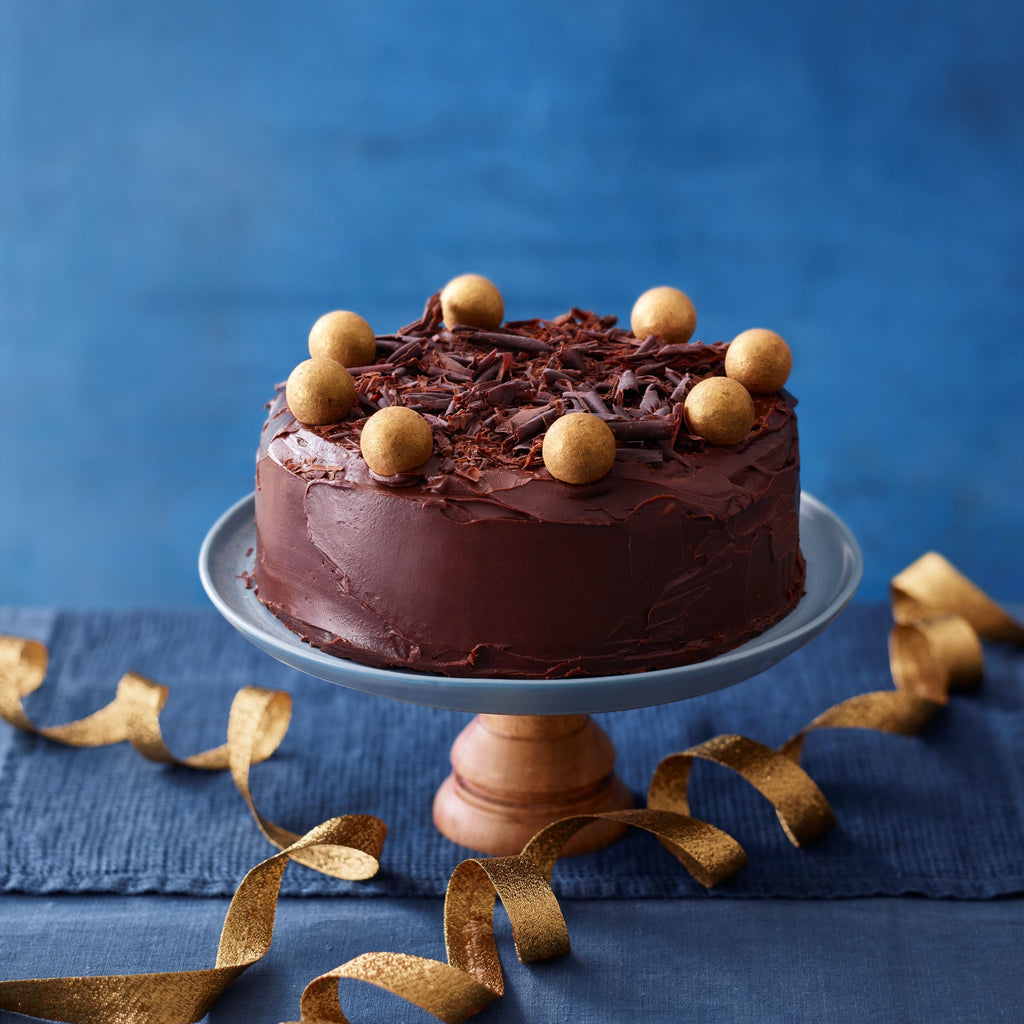Chocolate Fudge cake on a glass stand, decorated with gold chocolate truffles and chocolate shavings, styled with gold ribbon on a dark blue background.