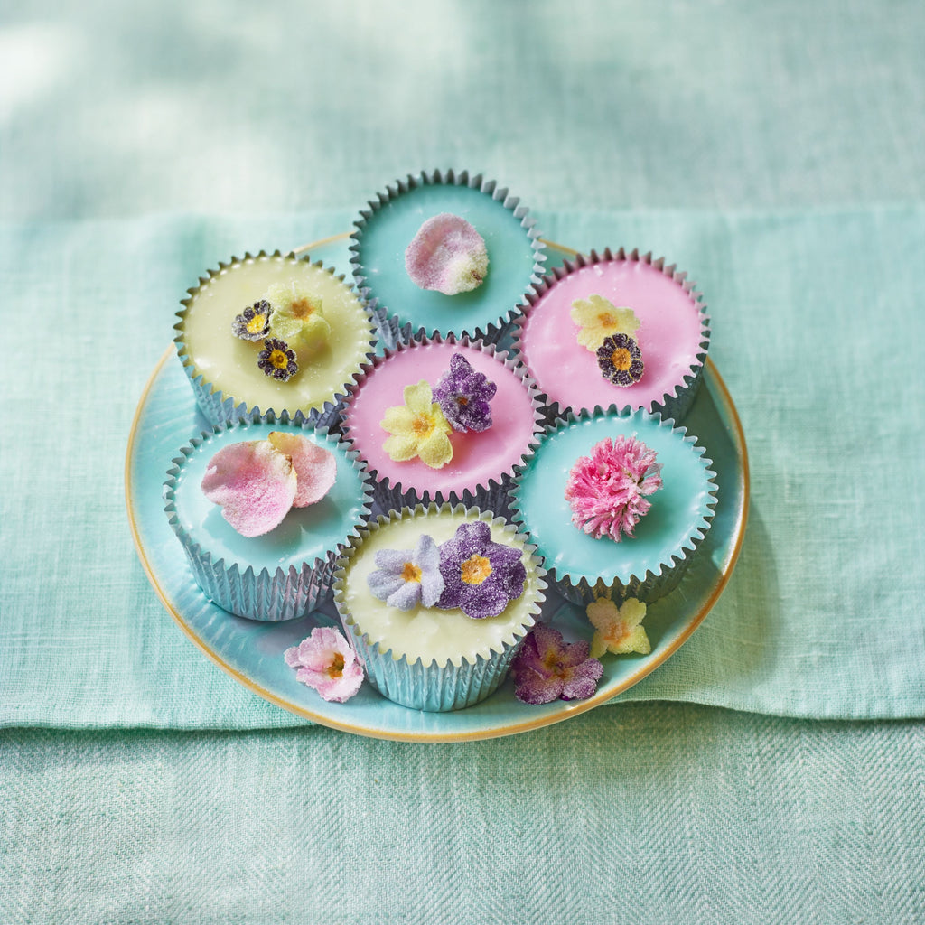 Seven pastel cupcakes topped with crystallised edible flowers on a turquoise plate, styled on a light green linen background.