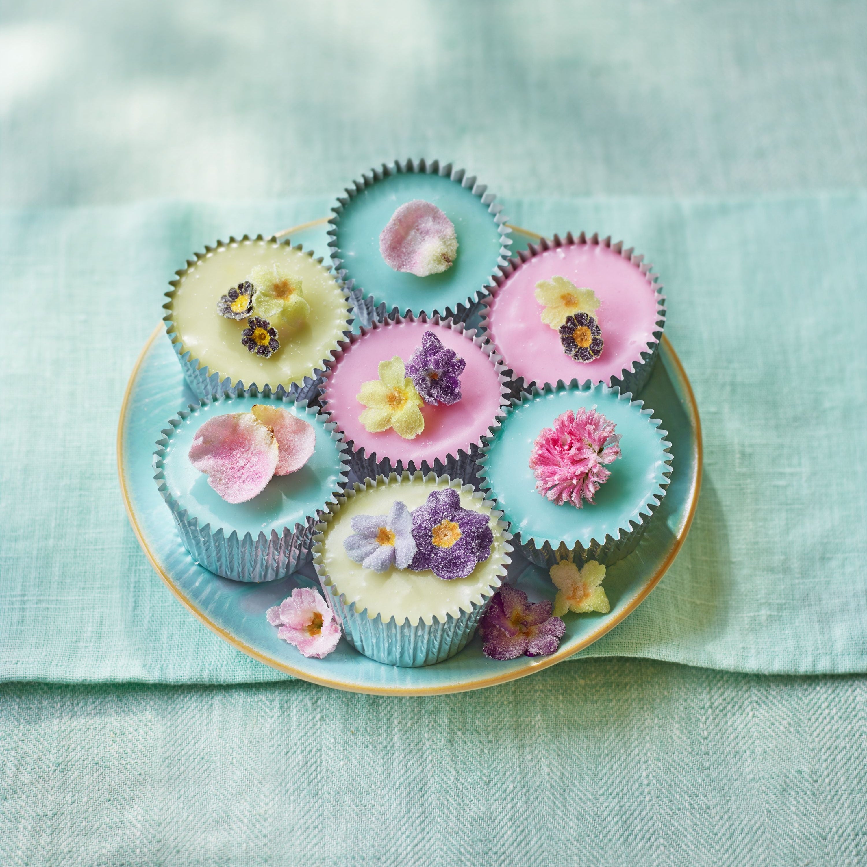 Seven pastel cupcakes topped with crystallised edible flowers on a turquoise plate, styled on a light green linen background.
