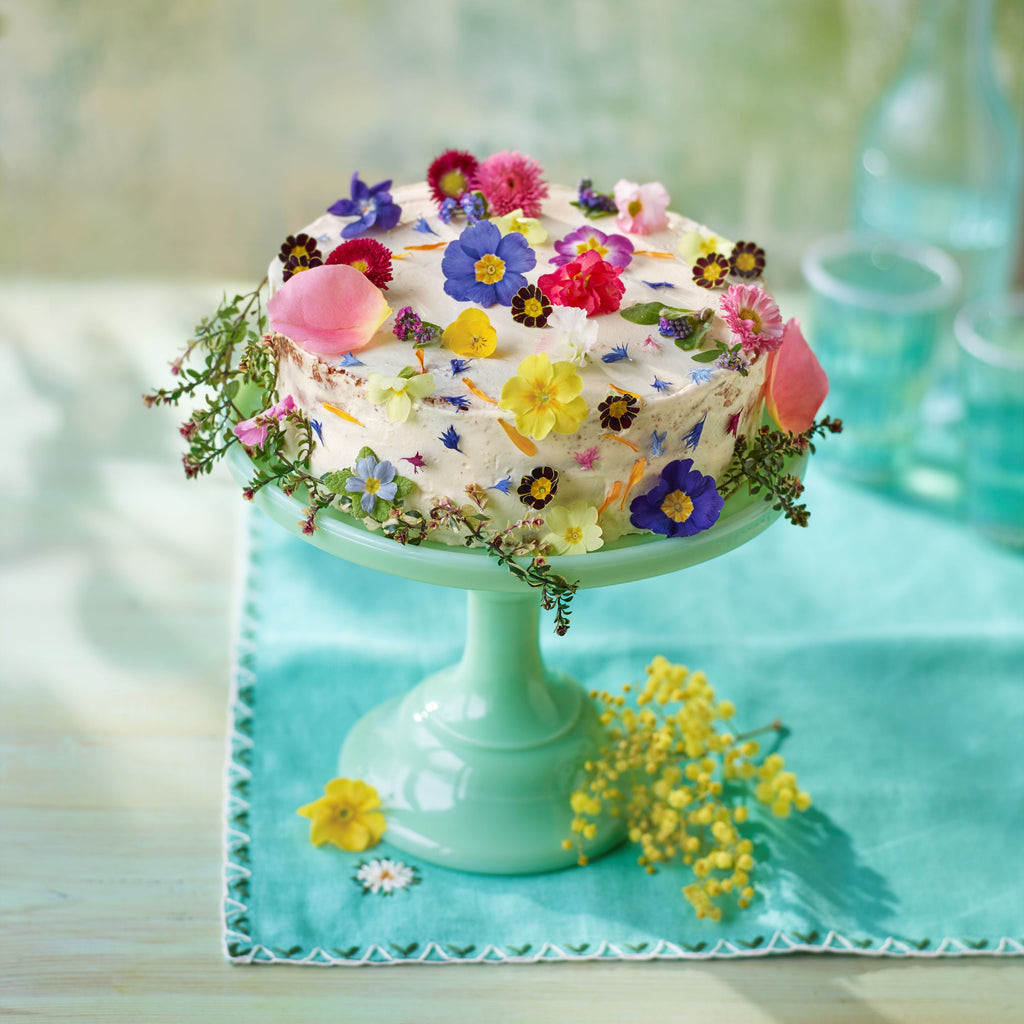 Buttercream cake decorated with colourful edible flowers on a mint green cake stand, set on a turquoise cloth with glass bottles in the background.