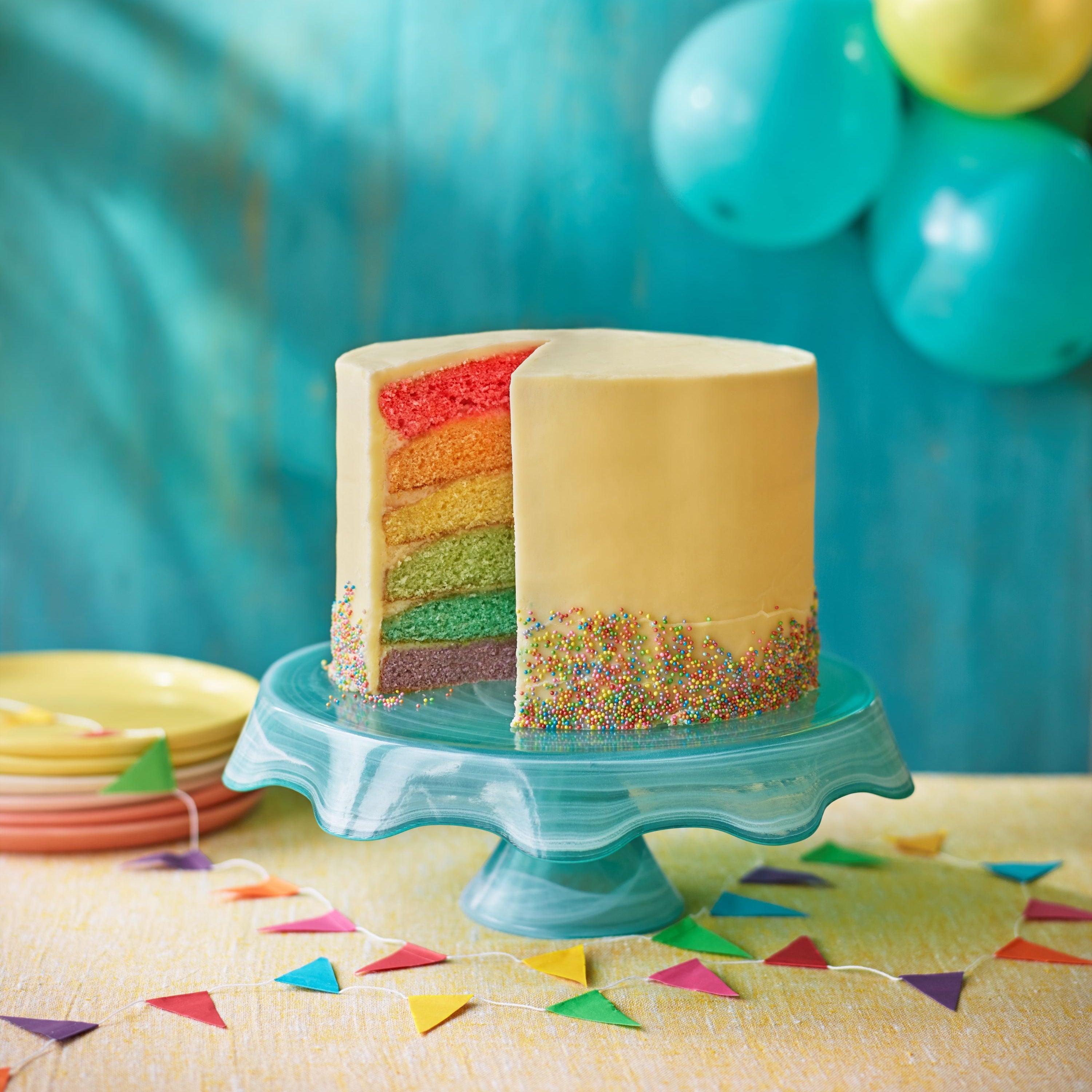 A rainbow layer cake with yellow frosting and sprinkles on a blue cake stand, surrounded by colourful balloons and bunting decorations on a party table.