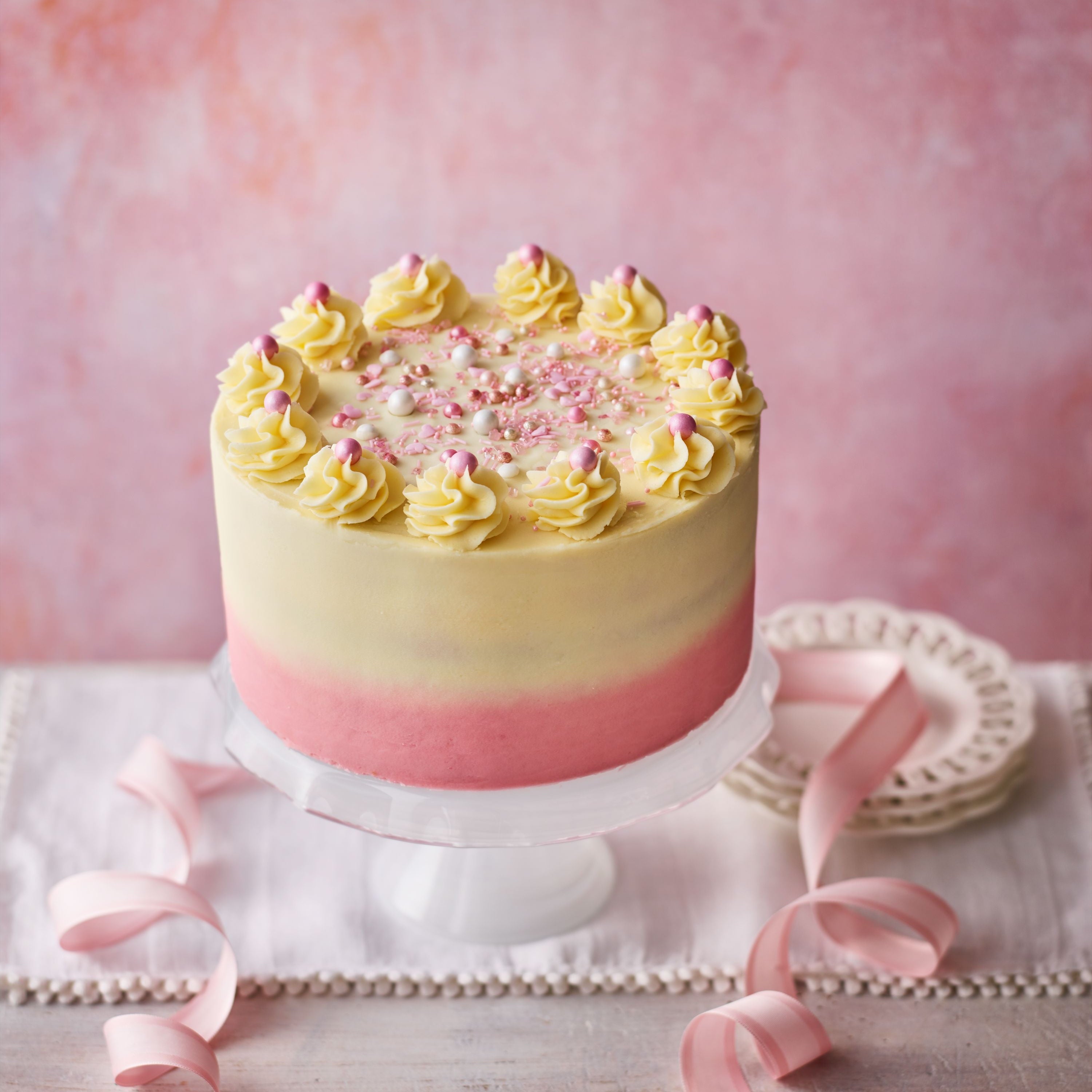 A pink and yellow ombré buttercream cake with swirl piping and sprinkles, styled with pink ribbon for birthdays and celebrations.