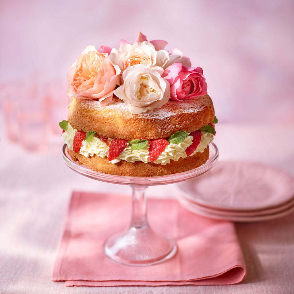 Strawberries and cream sponge cake decorated with fresh strawberries and roses on a glass stand, photographed on a soft pink background.